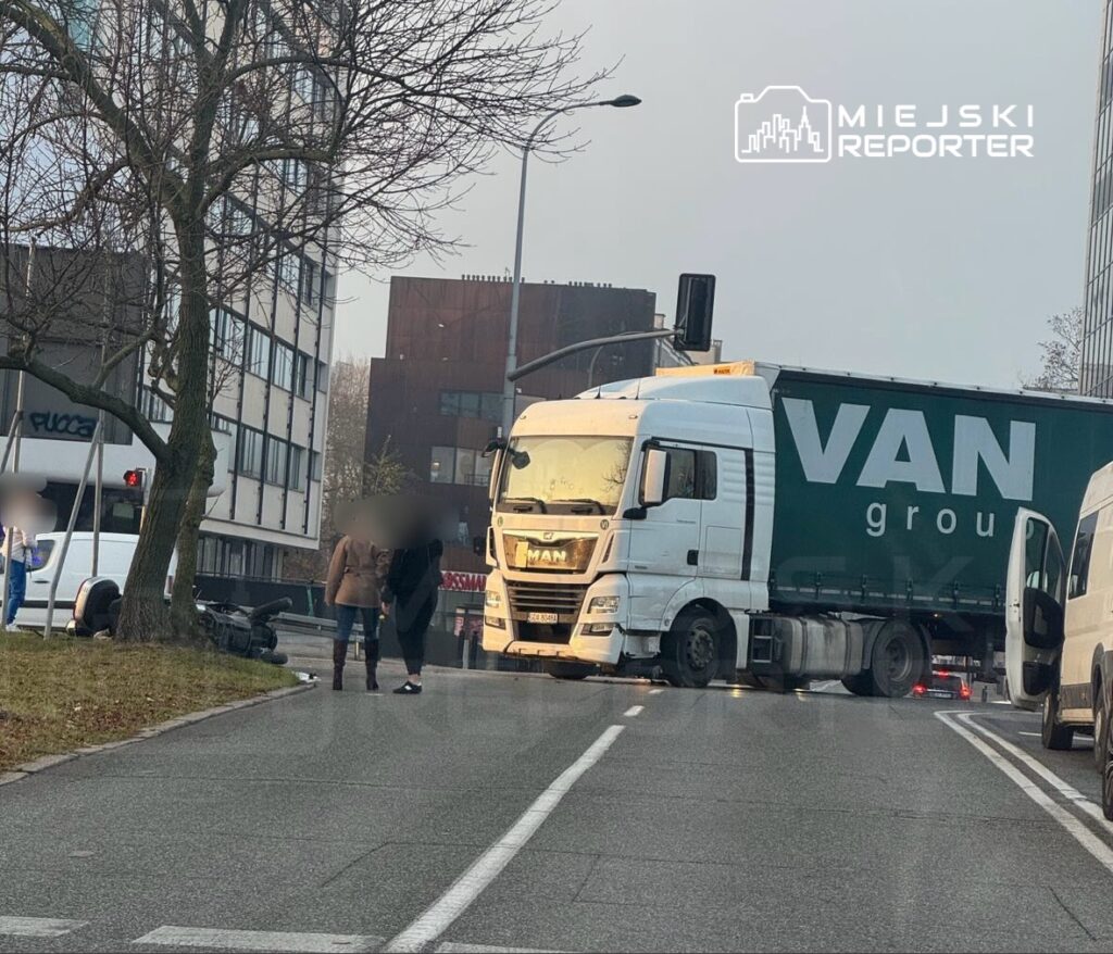 Czarny motocykl leży na boku, obok dwóch osób rozmawiających z kierowcą ciężarówki z logo "VAN group" na ruchliwej ulicy.