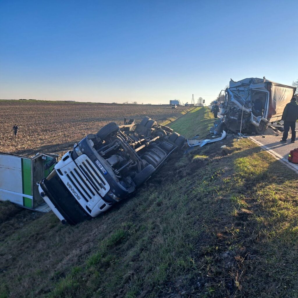 Czarny ciężarówka leży na boku w rowie, obok uszkodzona naczepa, a w tle widoczni ratownicy na wiejskiej drodze.