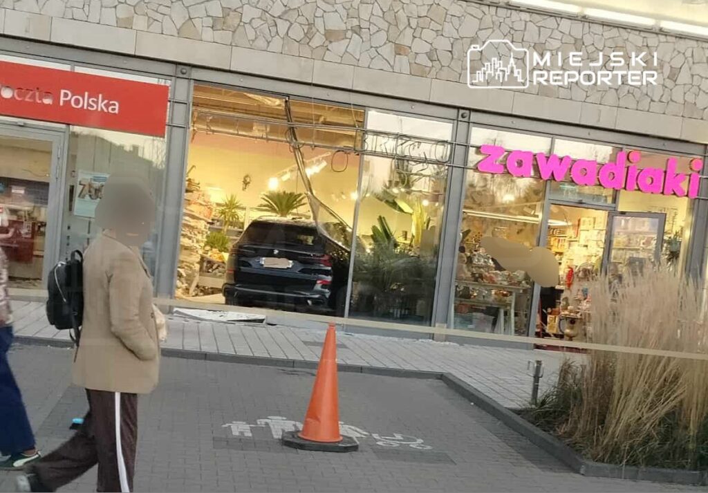 Mężczyzna w jasnym płaszczu spacerujący obok sklepu z różowym szyldem "zawadiaki" i samochodem w witrynie.