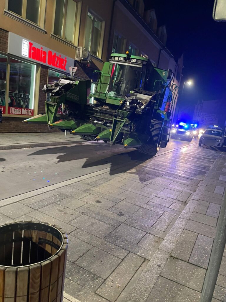 Zielony traktorek rolniczy stoi na ulicy w nocy, obok sklepu odzieżowego, w tle migają niebieskie światła radiowozu.