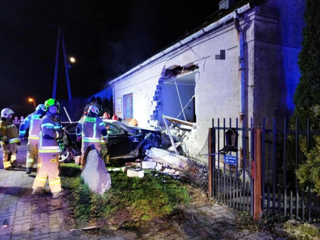Grupa strażaków pracuje przy zniszczonym budynku, w który uderzył samochód, nocą w oświetlonym miejscu.