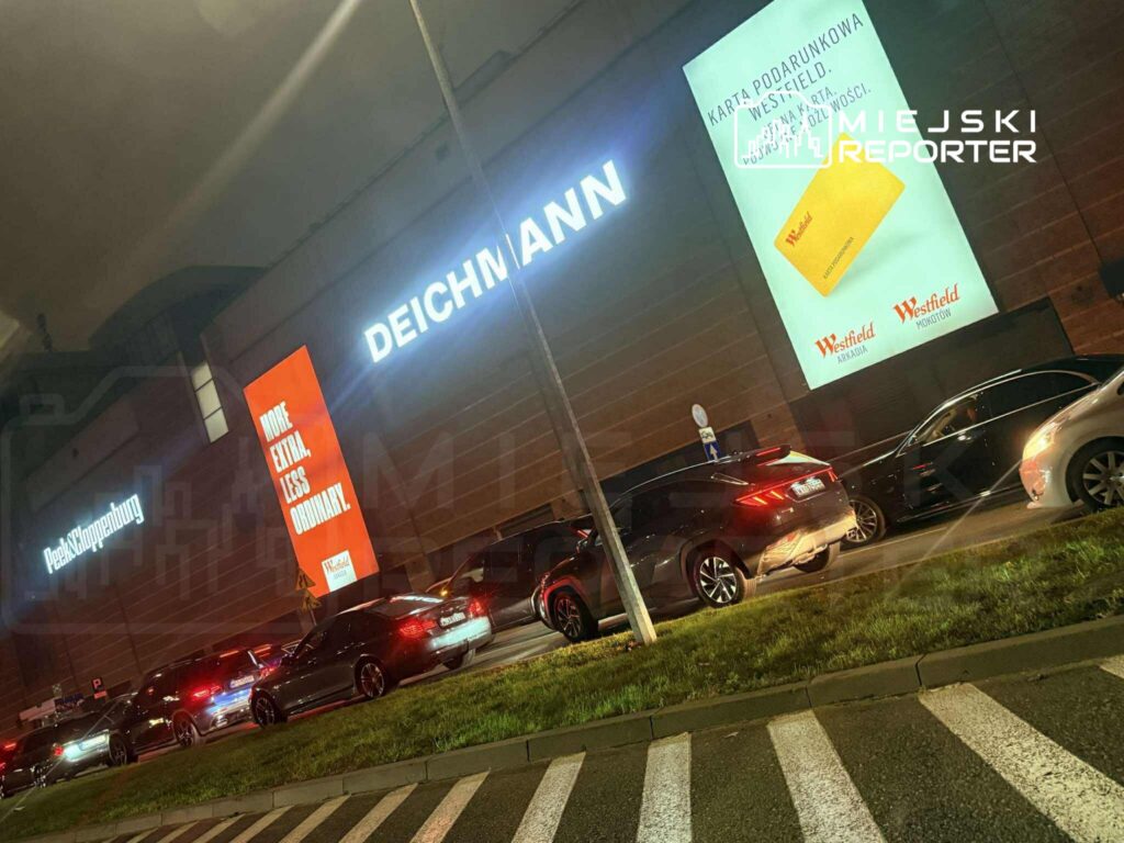 Ruch samochodowy przed nowoczesnym centrum handlowym, z widocznymi reklamami Deichmanna i Westfield.
