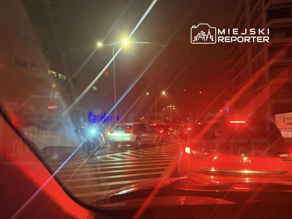 Ruch samochodowy w nocy na ulicy przed marketem, z widocznymi światłami pojazdów i znakami drogowymi.