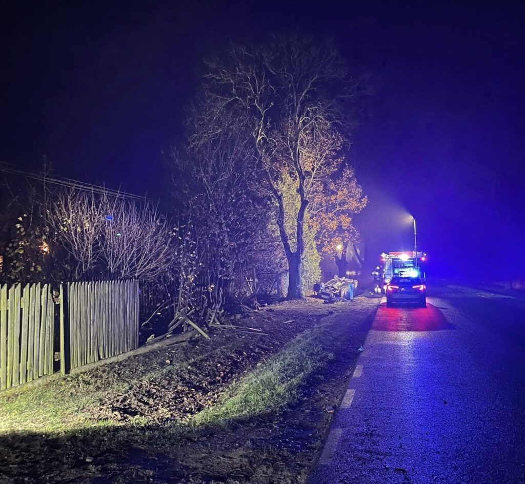 Na drodze nocą, nieoznakowany samochód policyjny oraz woz strażacki z włączonymi sygnalizatorami, w tle oświetlone drzewa.