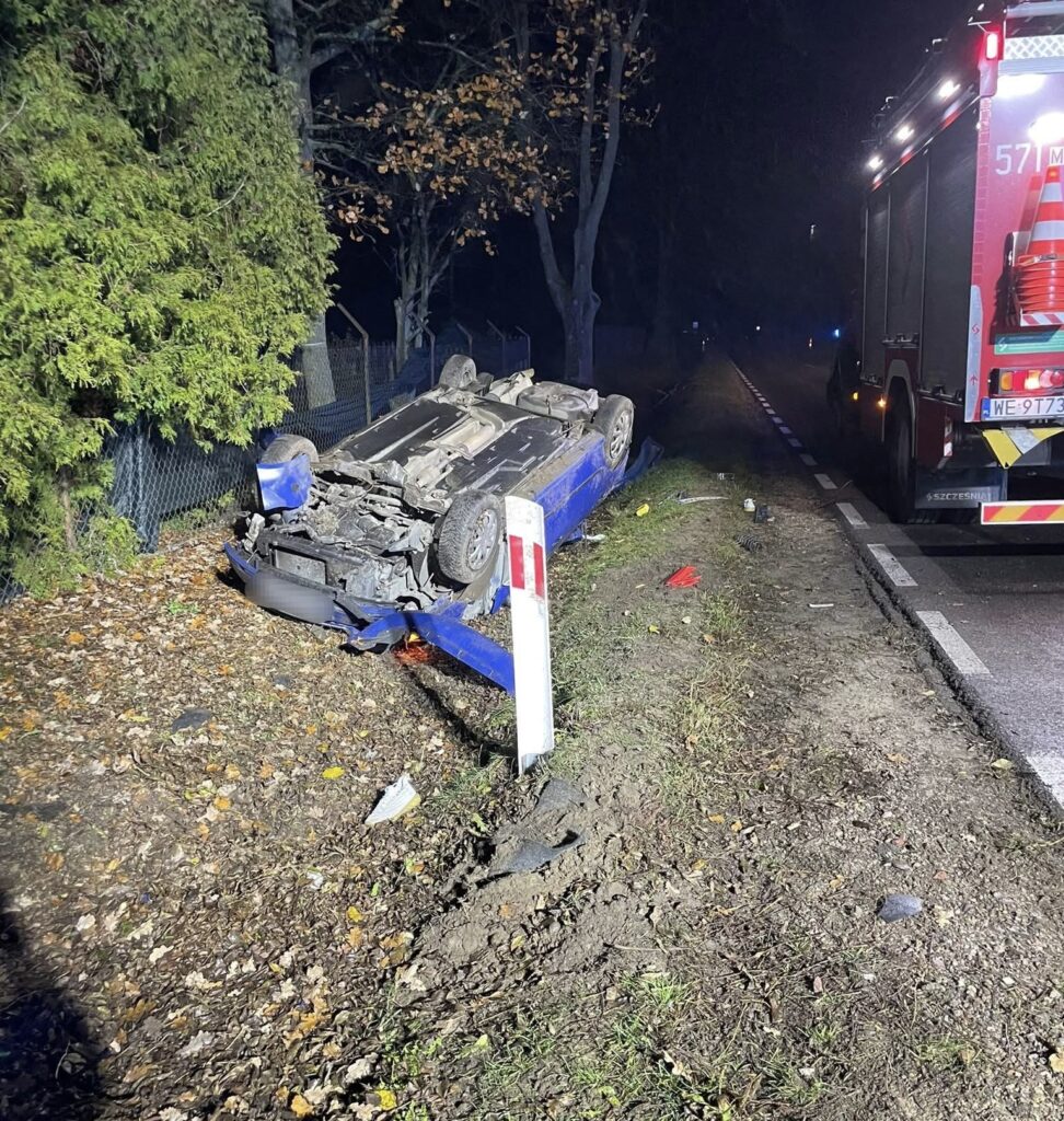 Przewrócone niebieskie auto leży na poboczu drogi, obok wozu strażackiego, w otoczeniu drzew i liści.