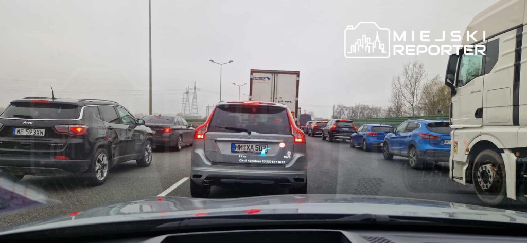 Ruch samochodowy w korku na drodze, widoczne różne pojazdy, w tym SUV-y i ciężarówki, w szarym, deszczowym otoczeniu.