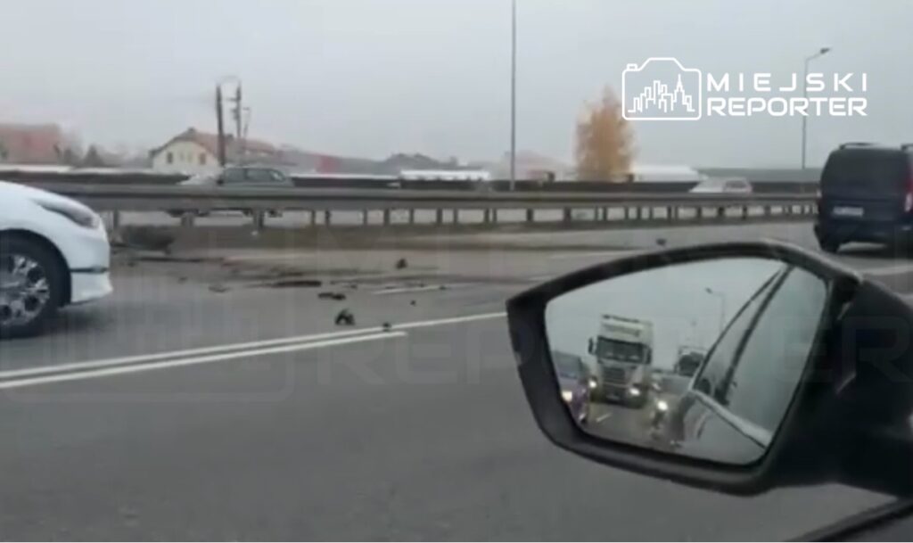 Na drodze widać uszkodzone pojazdy oraz pozostałości po wypadku, a w tle poruszające się samochody.