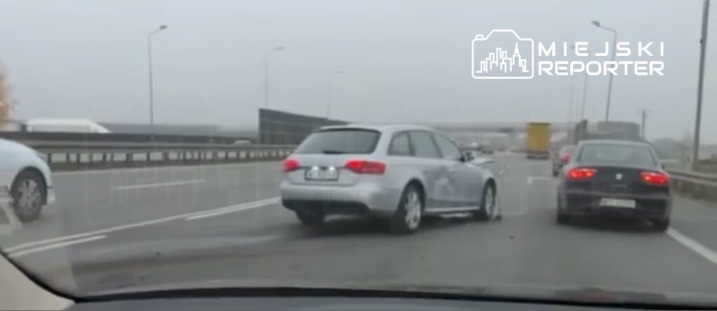 Srebrny samochód osobowy zmienia pas ruchu na zatłoczonej drodze w deszczową pogodę. W tle widać inne pojazdy.