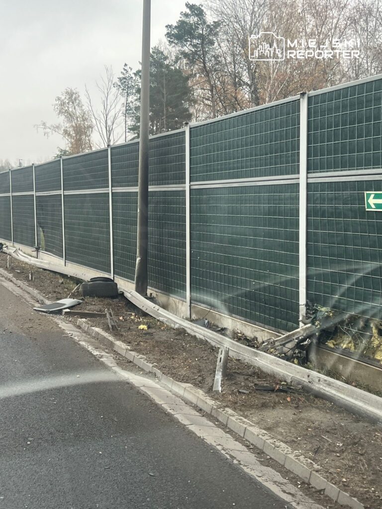 Uszkodzony fragment ogrodzenia dźwiękochłonnego obok drogi, z leżącymi resztkami opon i metalowymi elementami.