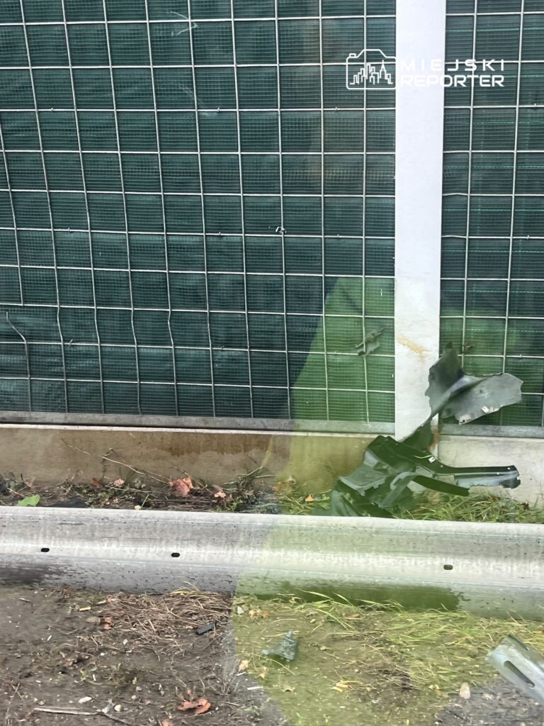 Zniszczona część metalowa leży na ziemi obok ogrodzenia z siatki, w tle widoczne zarośla.