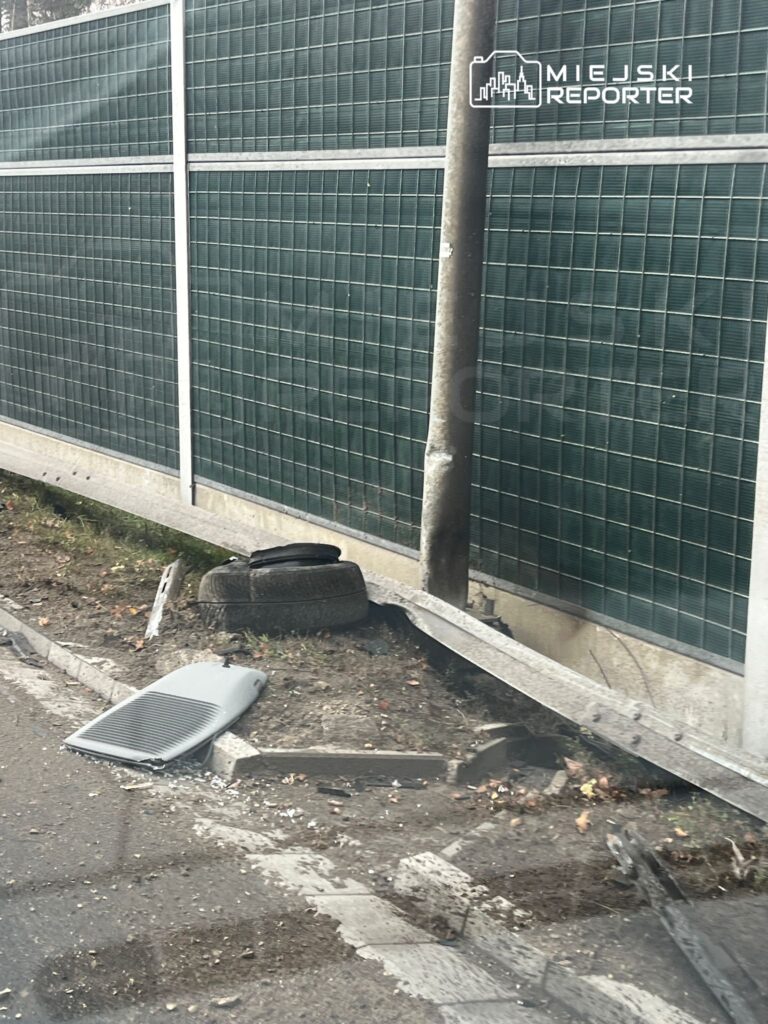 Zniszczona opona i fragment obudowy leżą na poboczu drogi obok metalowego płotu.