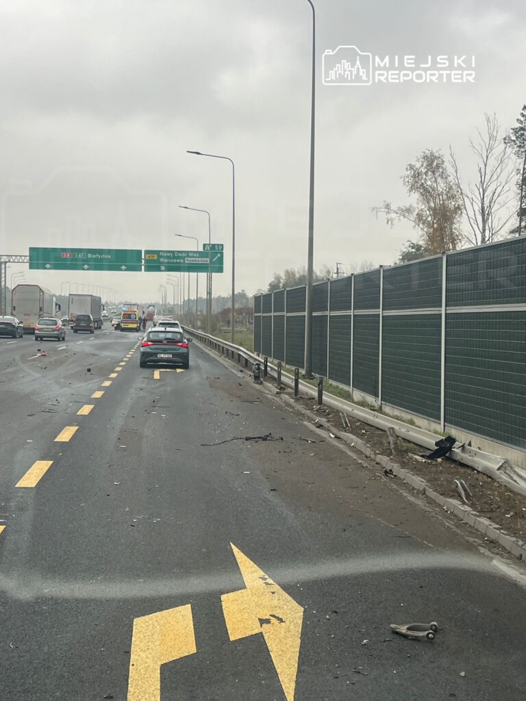 Na drodze widać uszkodzone pojazdy oraz rozbite elementy, obok przejeżdża ambulans. W tle znaki drogowe wskazujące kierunki.