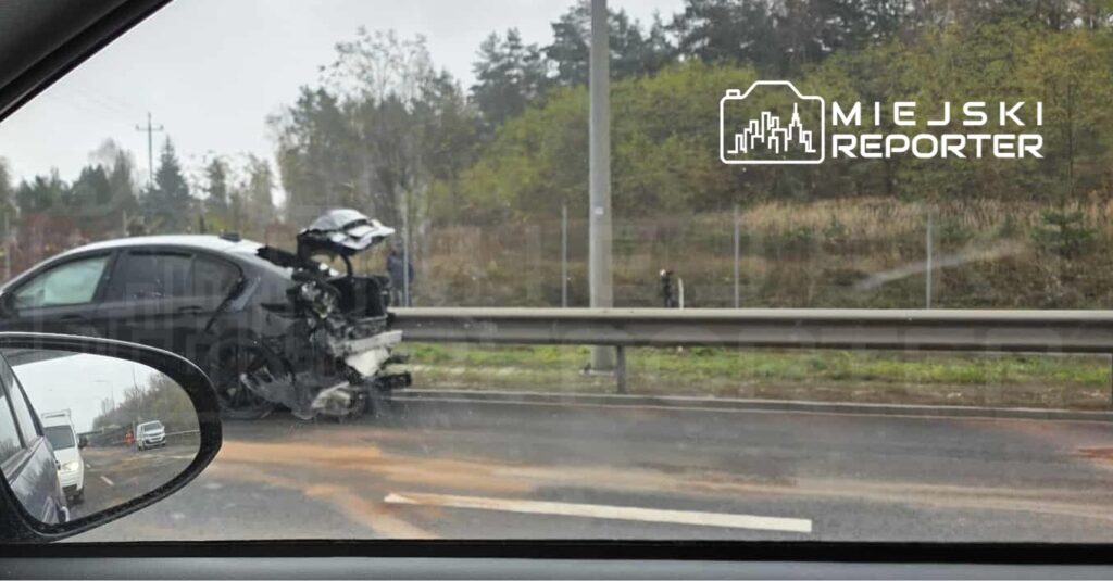 Uszkodzony samochód osobowy stoi przy barierce na poboczu drogi, obok widoczne inne pojazdy w ruchu.