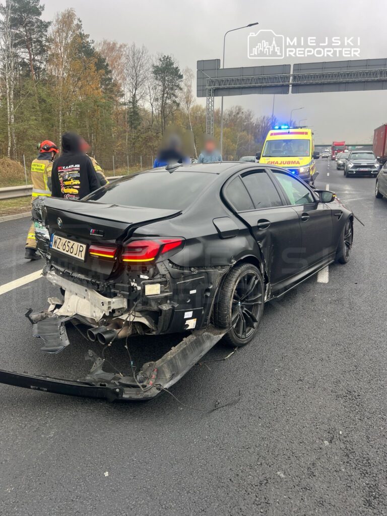 Uszkodzony czarny samochód osobowy na drodze, obok grupy osób oraz pojazdu z sygnałami świetlnymi.