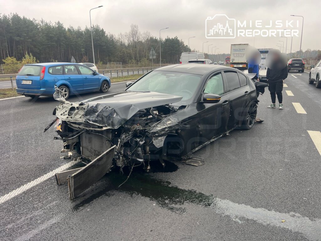 Czarny samochód osobowy z uszkodzoną przednią częścią stoi na drodze, obok dwóch osób rozmawiających.