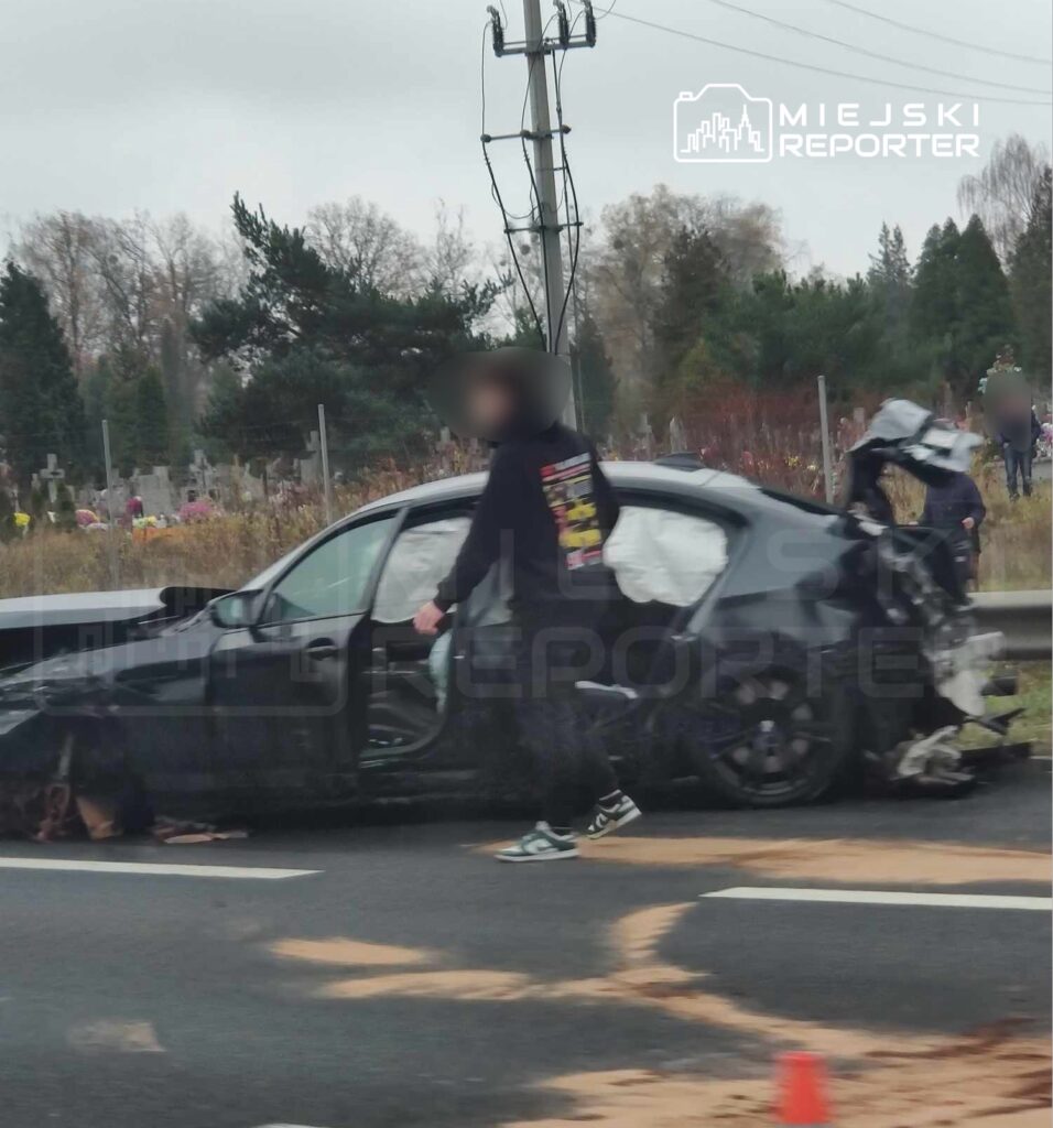 Mężczyzna w czarnej bluzie z kapturem sprawdza uszkodzone auto na poboczu drogi obok cmentarza.