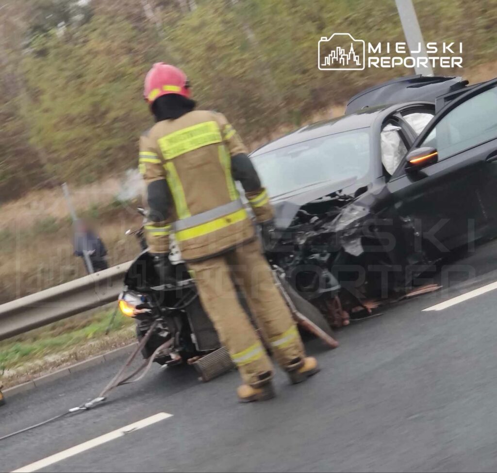 Strażak w mundurze z kaskiem ocenia uszkodzenia czołowo zderzonego samochodu na drodze.