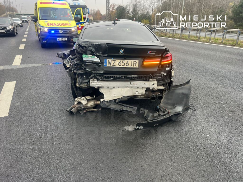 Uszkodzony czarny samochód osobowy stoi na drodze, a w tle widać ambulans z włączonymi sygnałami świetlnymi.