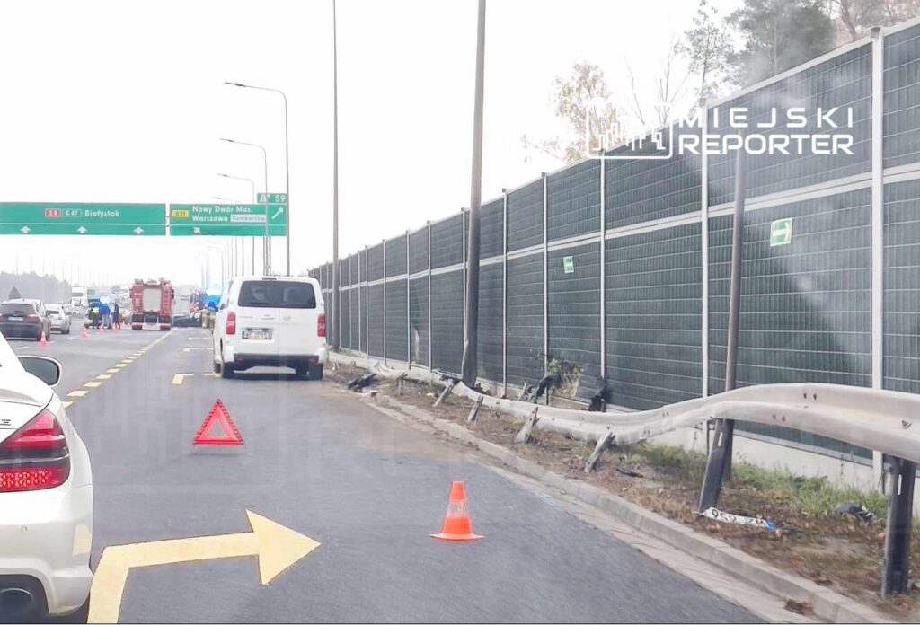 Na jezdni stoi biały samochód dostawczy, a obok widać woz strażacki i kilka osób w mundurach. W tle znaki drogowe wskazujące kierunki.