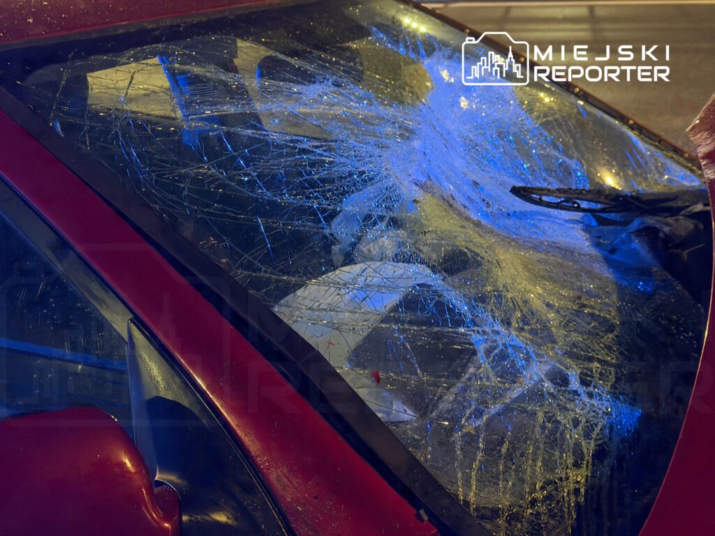Zniszczona przednia szyba czerwonego samochodu z widocznymi pęknięciami, oświetlona niebieskim światłem.