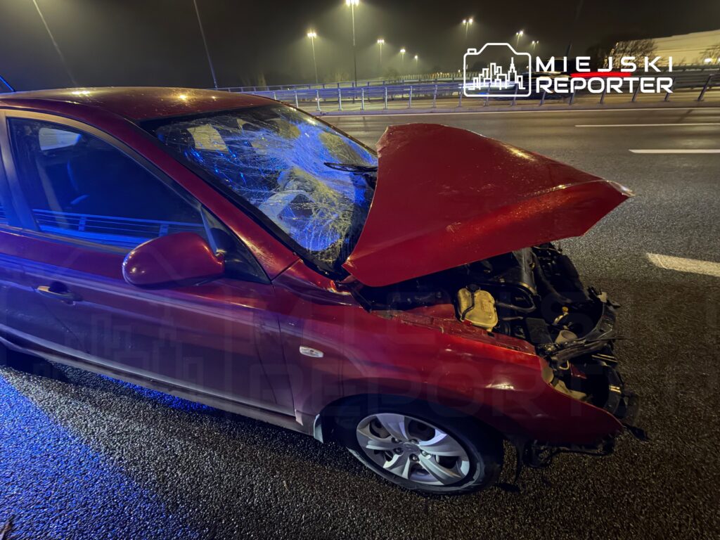 Czerwony samochód osobowy z uszkodzoną maską i pękniętą szybą stoi na drodze w nocy, otoczony niebieskim światłem.