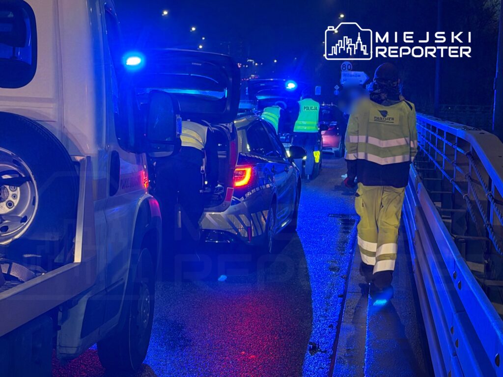 Funkcjonariusze policji oraz straży pożarnej prowadzą akcję na oświetlonej drodze, obok zatrzymanego pojazdu.