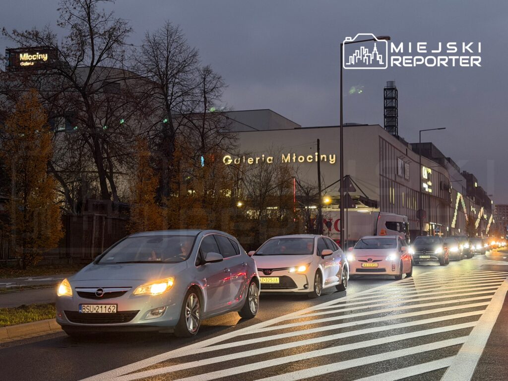 Ruch samochodowy na pasie drogowym przed Galerią Młociny w wieczornej scenerii.