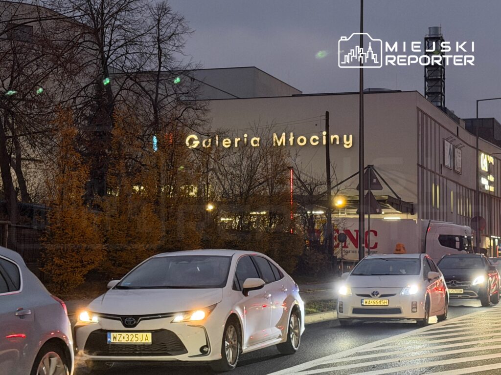 Ruchliwe skrzyżowanie z samochodami w kolejce, w tle widoczny neon "Galeria Młociny" w wieczornym świetle.