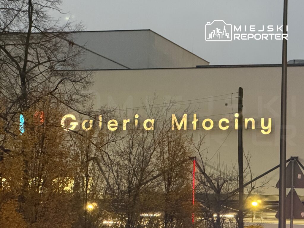 Podświetlony napis "Galeria Młociny" na tle wieczornego nieba i drzew w okolicy.