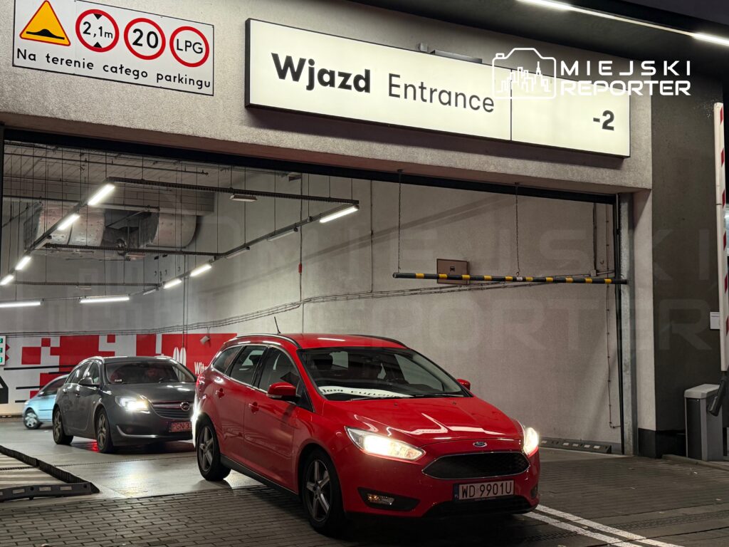 Czerwony samochód wjeżdża do parkingu podziemnego, obok stoi szary pojazd, w tle widoczna tablica z napisem "Wjazd".