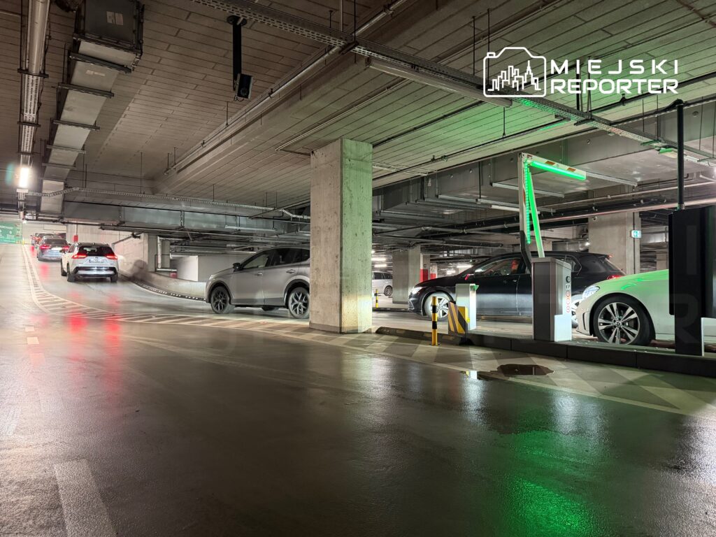 Ruch samochodów w podziemnym parkingu z oświetleniem LED i mokrą nawierzchnią, w tle widoczne wyjazdy.