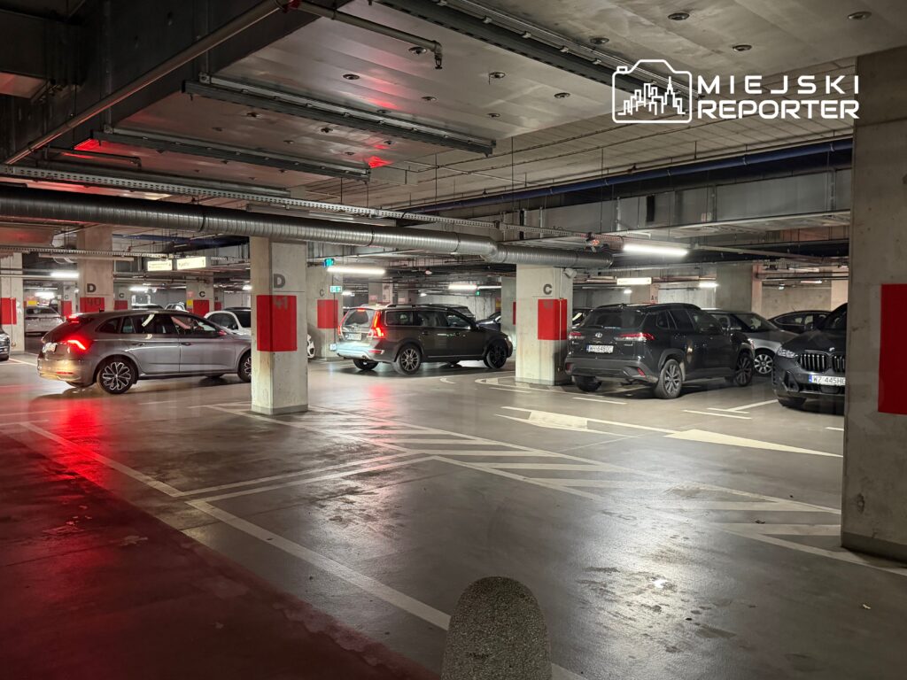 Parking podziemny z kilkoma samochodami, oświetlony sztucznym światłem, z oznaczeniami miejsc parkingowych.