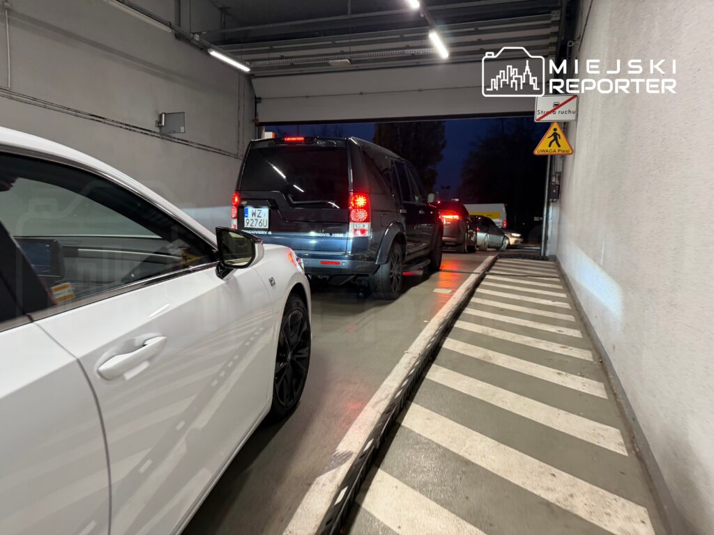 Ruch samochodowy w wąskim tunelu, z białym autem na pierwszym planie i ciemnym SUV-em w tle. Widać oznakowanie dla pieszych.