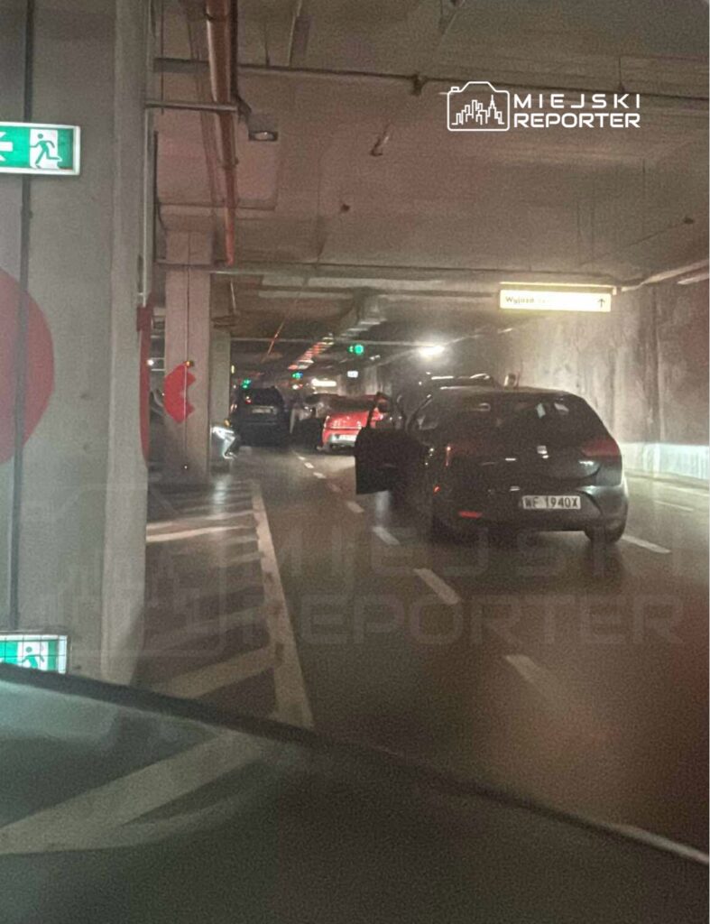 Czarny samochód zatrzymany w podziemnym parkingu, z otwartymi drzwiami, obok innych pojazdów. W tle widoczna zielona sygnalizacja.