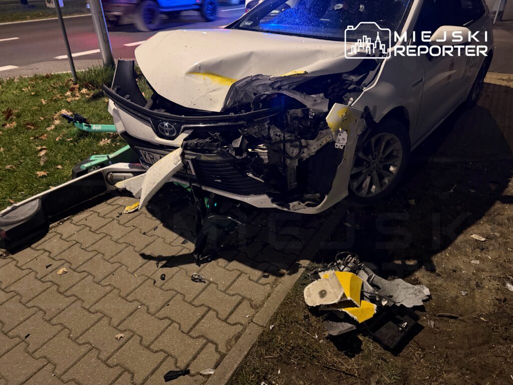 Rozbity samochód osobowy marki Toyota z uszkodzoną przednią częścią stoi na chodniku, obok leżą fragmenty skuterów.