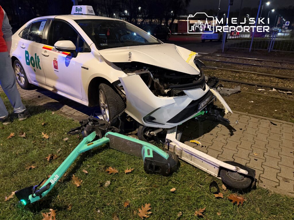 Zniszczony samochód taksówkowy z widocznym uszkodzeniem przodu, obok leży uszkodzony skuter elektryczny.