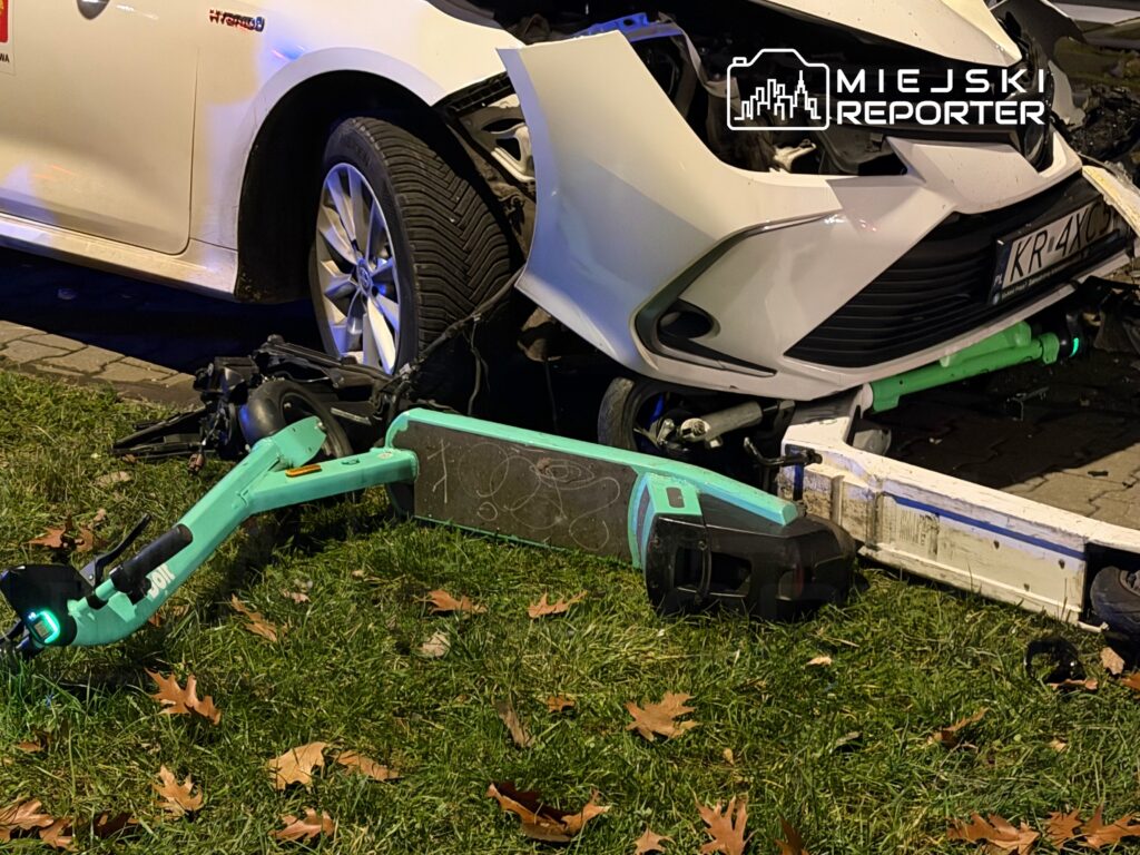 Zniszczona hulajnoga elektryczna leży na trawie obok uszkodzonego samochodu osobowego.