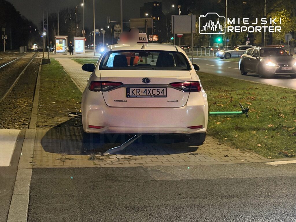 Białe taxi zaparkowane na chodniku obok torów tramwajowych, z zieloną hulajnogą opartą o tylną część pojazdu.