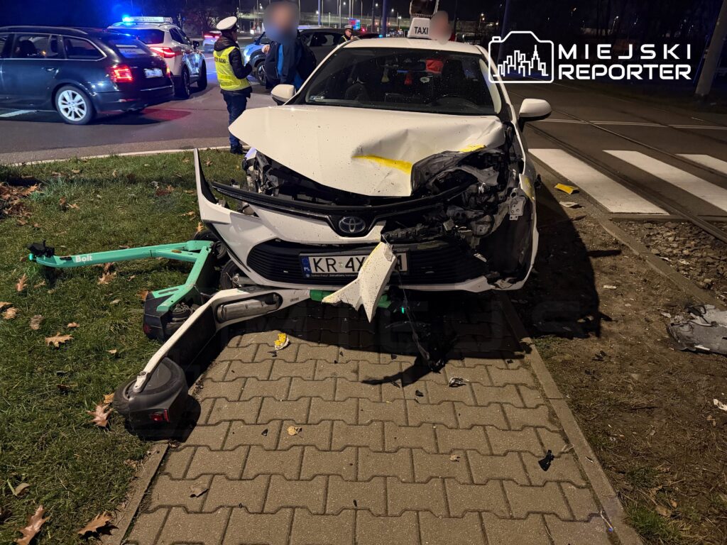 Rozbity taksówka marki Toyota stoi na chodniku, z uszkodzoną przednią częścią, obok policjant rozmawia z osobą.