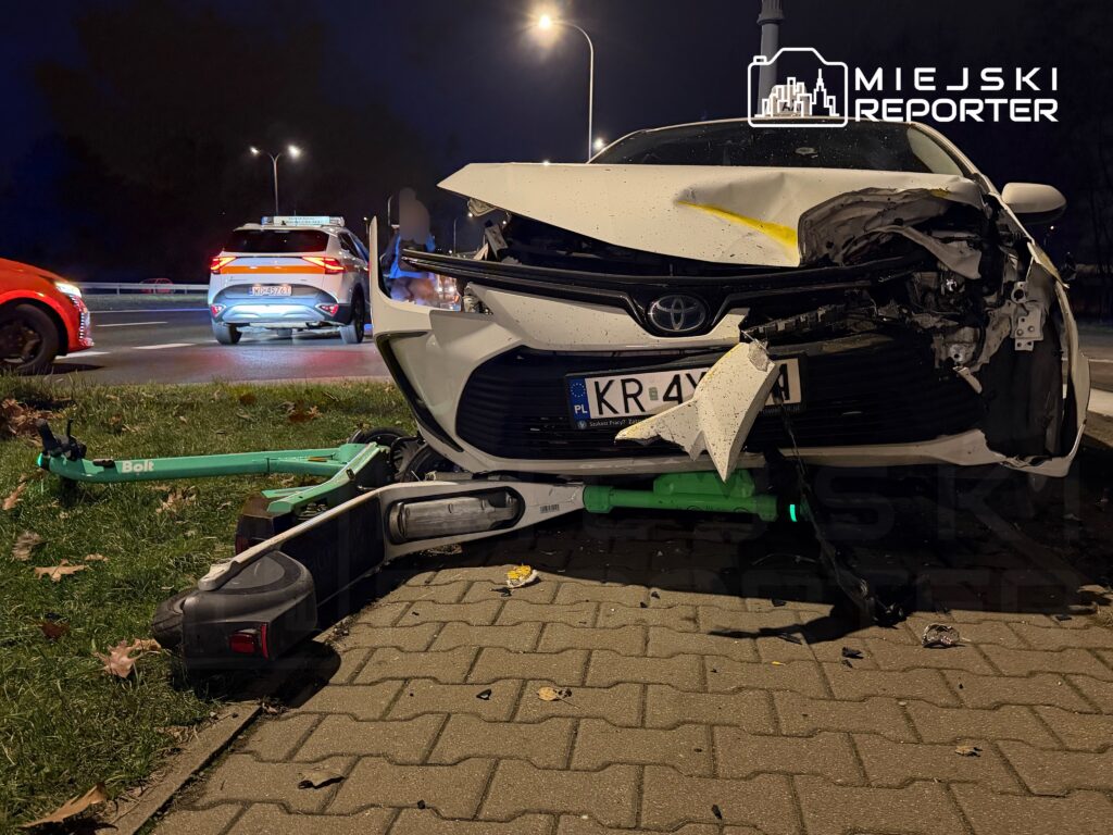 Zderzenie samochodu osobowego z uszkodzoną przednią częścią oraz leżącą na trawie hulajnogą elektryczną.