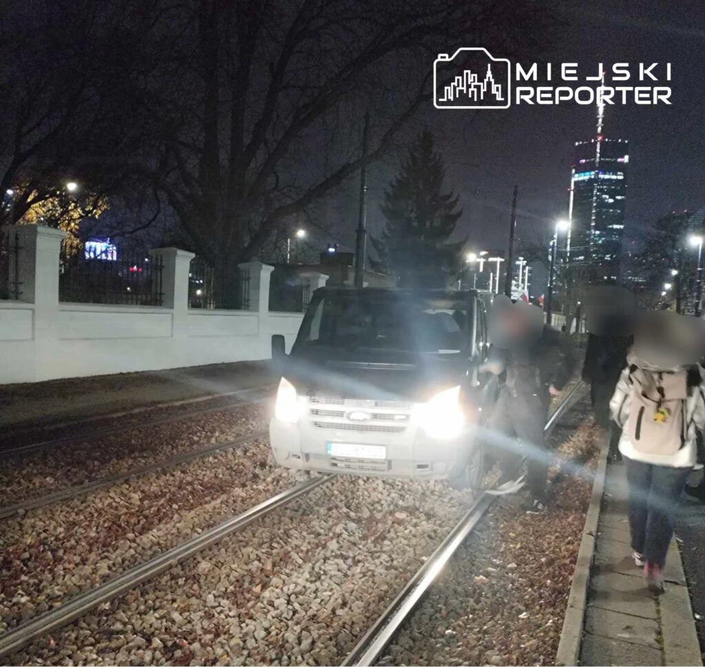 Samochód dostawczy stoi na torach tramwajowych, a obok grupka ludzi spaceruje wzdłuż chodnika nocą.