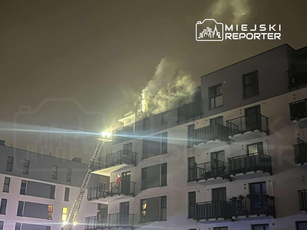 Strażacy używają drabiny do gaszenia pożaru na dachu nowoczesnego budynku mieszkalnego. W tle widać unoszący się dym.