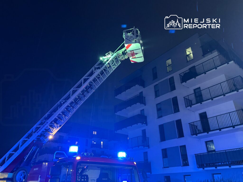 Straż pożarna używa drabiny, aby dotrzeć do okna na wyższym piętrze nowoczesnego budynku mieszkalnego w nocy.