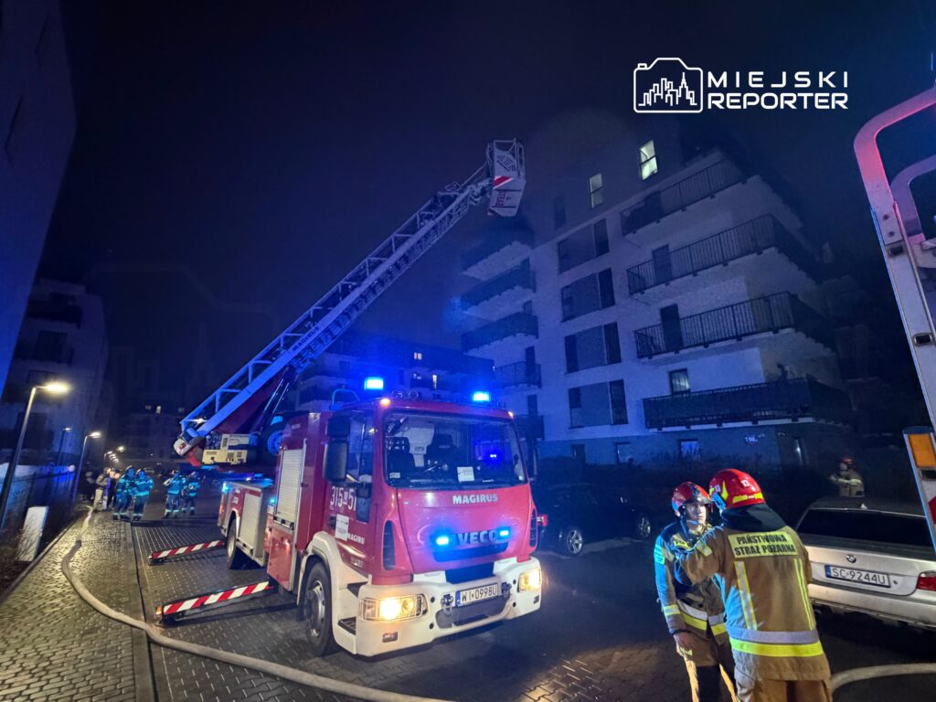 Strażacy w mundurach rozmawiają obok wozu strażackiego z podnośnikiem w nocy przy nowoczesnym budynku mieszkalnym.