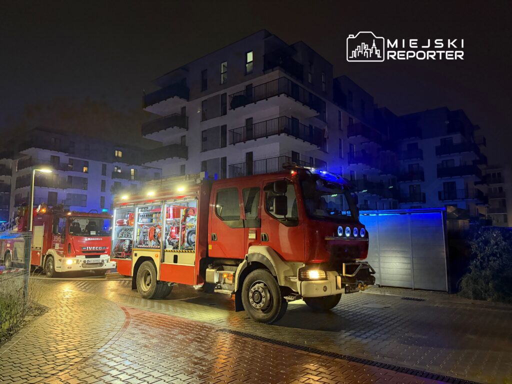 Czerwony wóz strażacki z włączonymi sygnałami świetlnymi stoi na oświetlonej ulicy przed nowoczesnym budynkiem mieszkalnym.