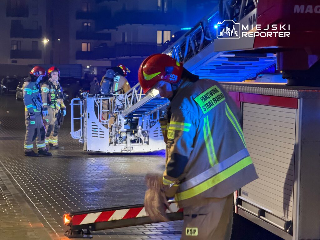 Grupa strażaków w hełmach i mundurach przygotowuje sprzęt przy wozie strażackim na oświetlonym placu.