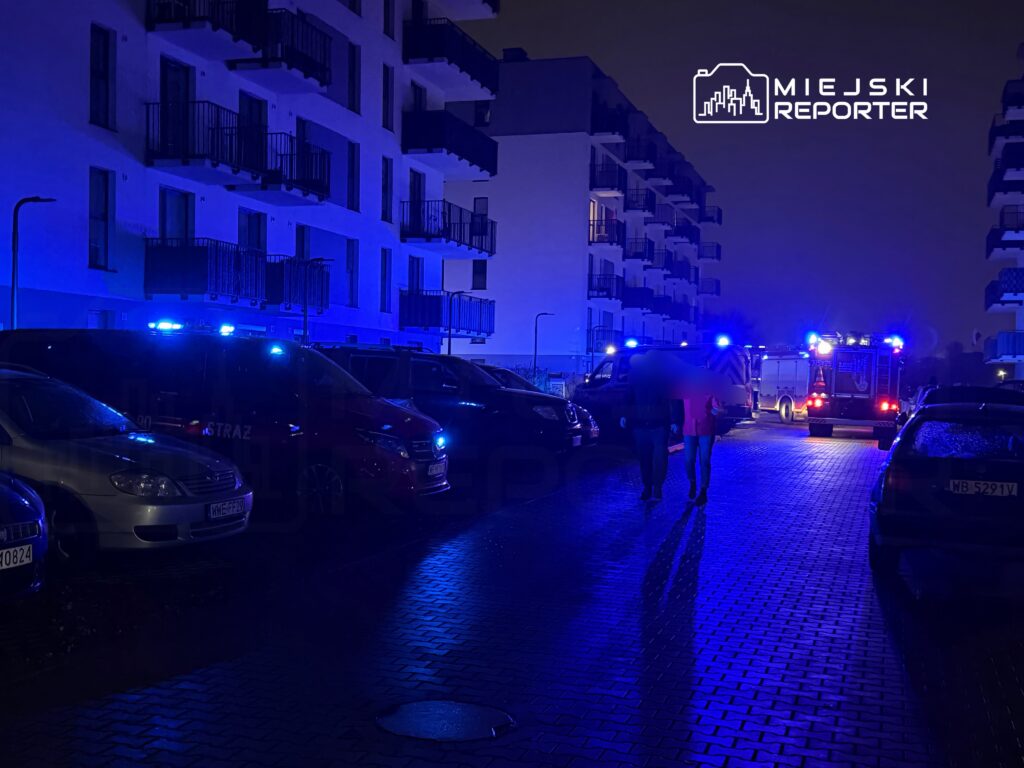 Dwóch ludzi spaceruje wzdłuż oświetlonej niebieskimi światłami ulicy, obok zaparkowanych samochodów, w tle widać pojazdy straży pożarnej.