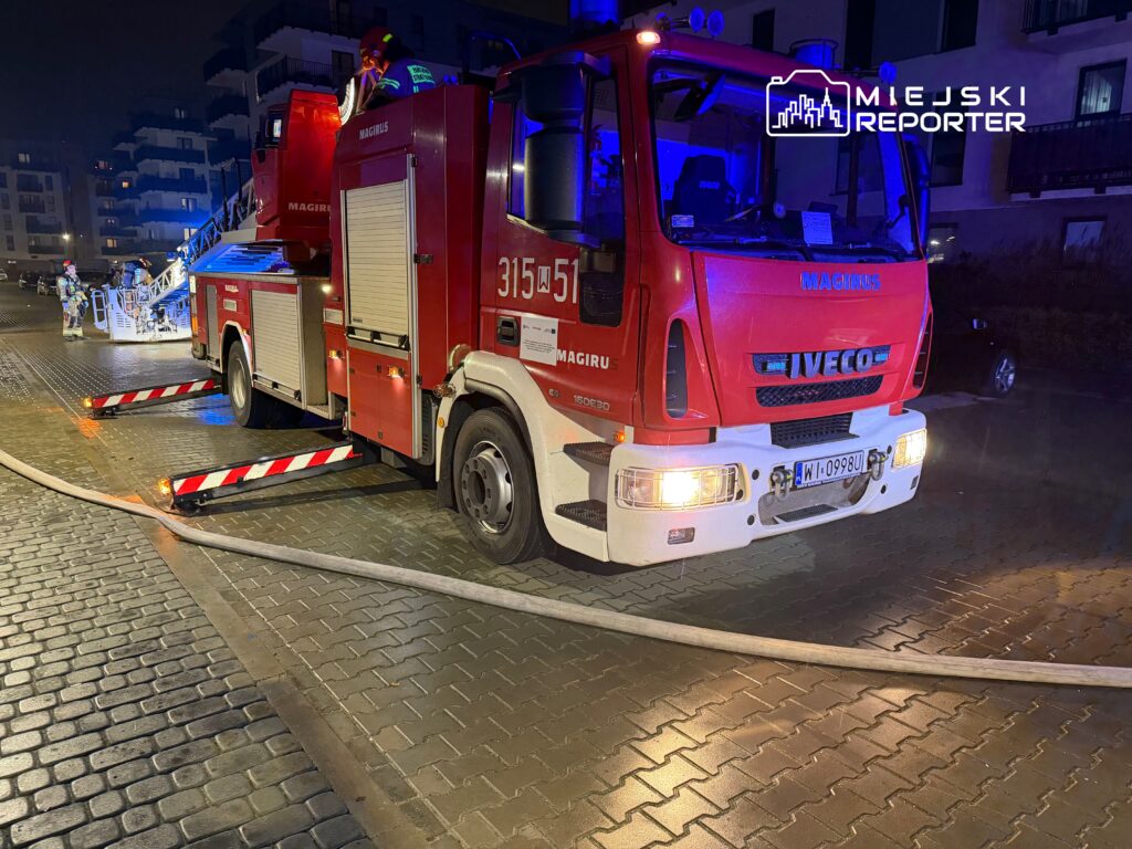 Czerwony wóz strażacki z drabiną i włączonymi sygnałami świetlnymi stoi na oświetlonej ulicy.