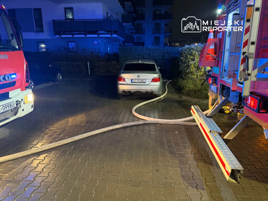 Straż pożarna podjeżdża do budynku mieszkalnego, rozkładając węże obok zaparkowanego samochodu.