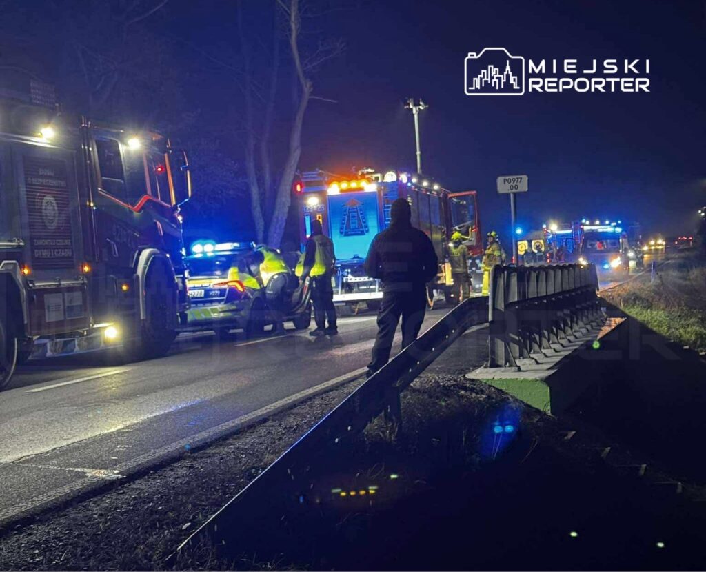 Ratownicy i policjanci pracują przy wypadku drogowym na ciemnej, oświetlonej ulicy, otoczeni przez pojazdy służb.